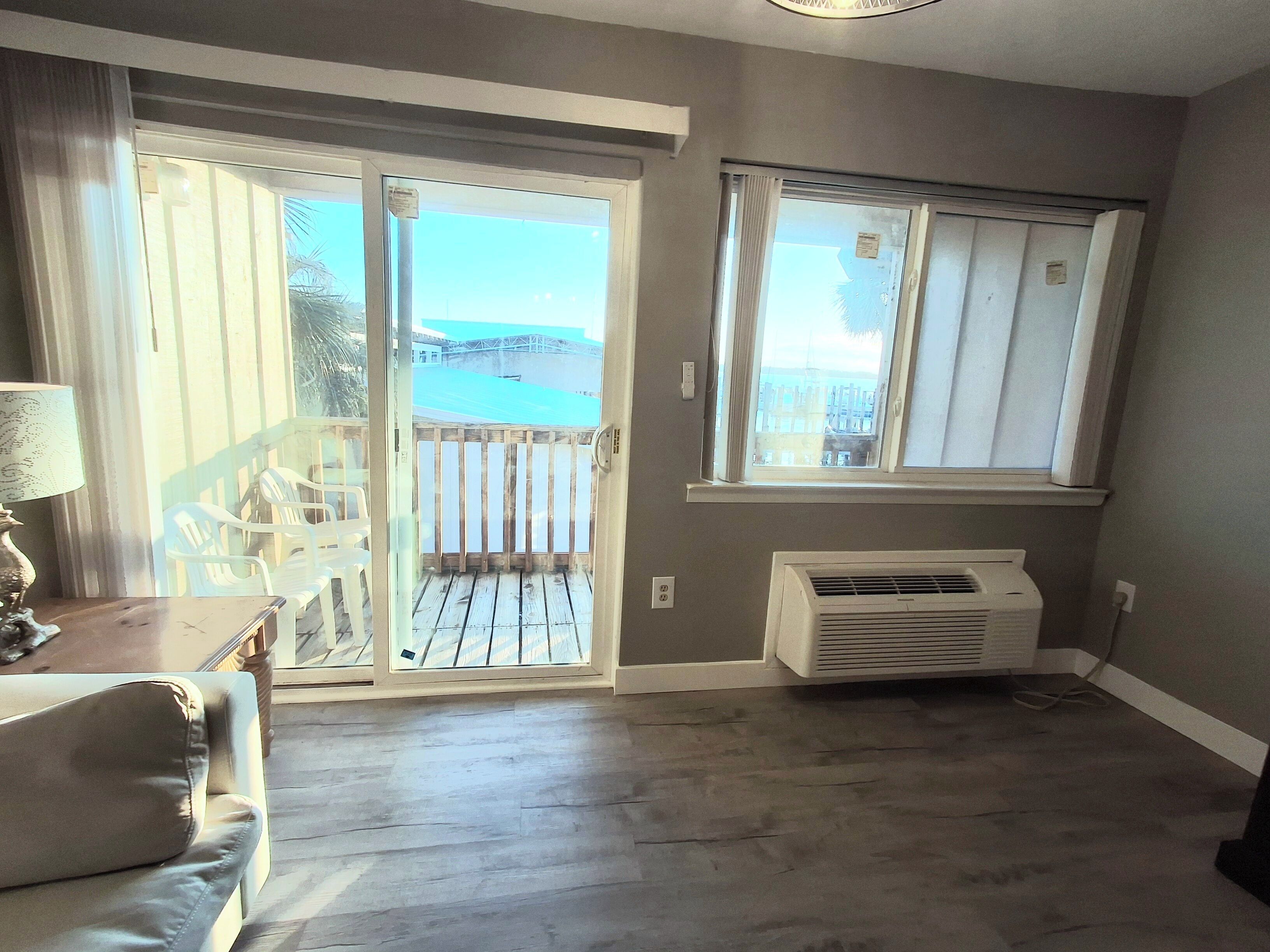 A room with a sliding glass door leading to a balcony, a white air conditioning unit, and a view of a building across the street.