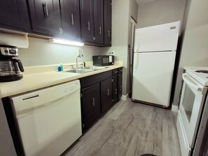A compact kitchen with dark cabinets, a white dishwasher, a white refrigerator, a microwave, and a coffee maker. - Sun Harbor Overlook - Sleeps 4 - 1 Bedroom Condo - Waterfront View (Panama City)