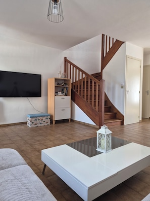 A living room with a flat-screen TV, a wooden staircase, a white coffee table, and a patterned ottoman. - Family Home in Groix – Sleeps 8 – Garden & Bikes (Groix)