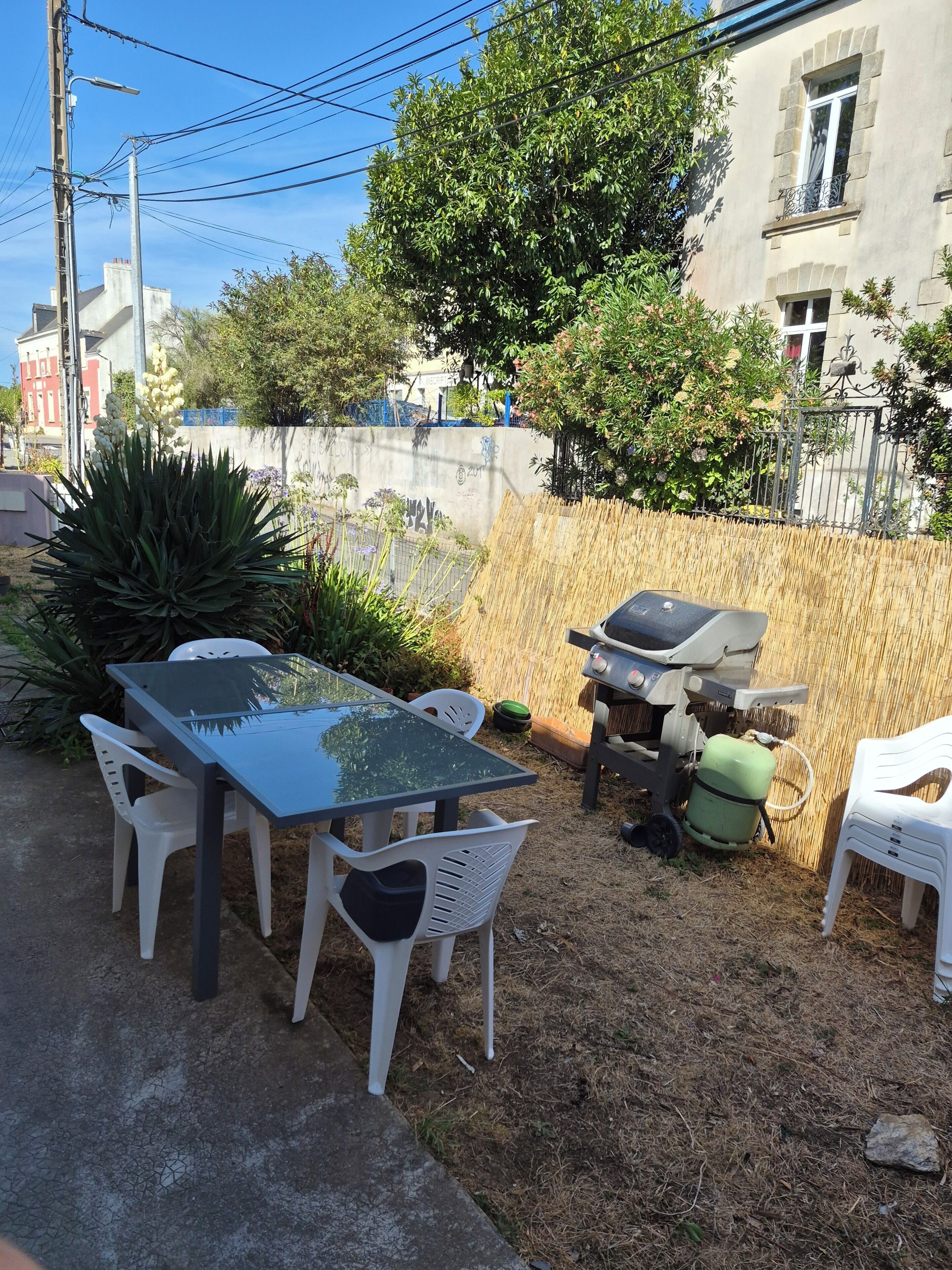 An outdoor patio with a glass table and white plastic chairs, surrounded by a bamboo fence and various plants.