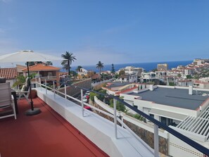 Ein Balkon mit rotem Boden, weißem Geländer und Blick auf eine Küstenstadt mit Gebäuden und Palmen.