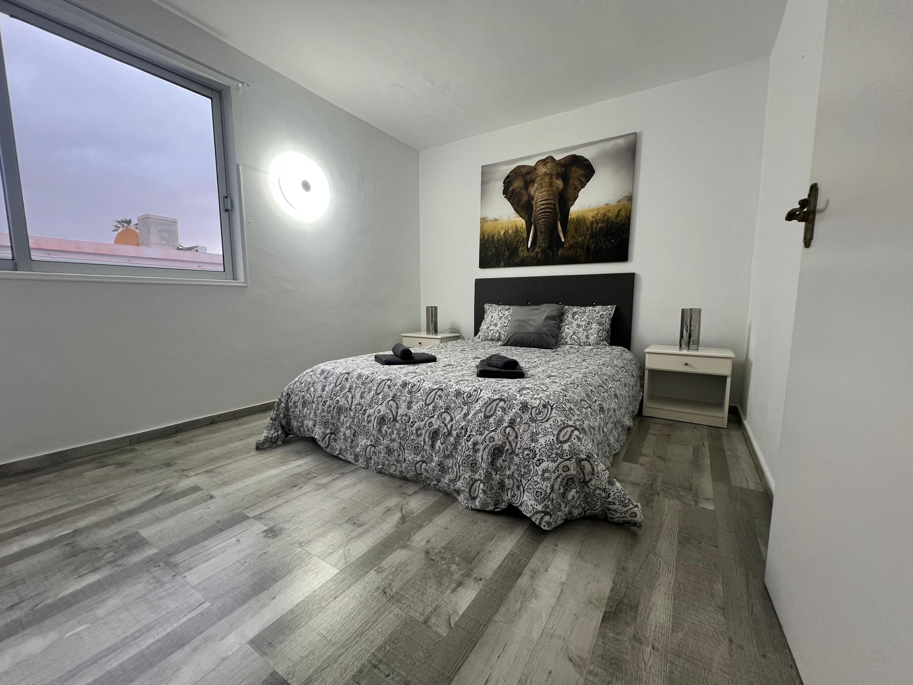A bedroom with a bed, a nightstand, a wall-mounted light, and a window.