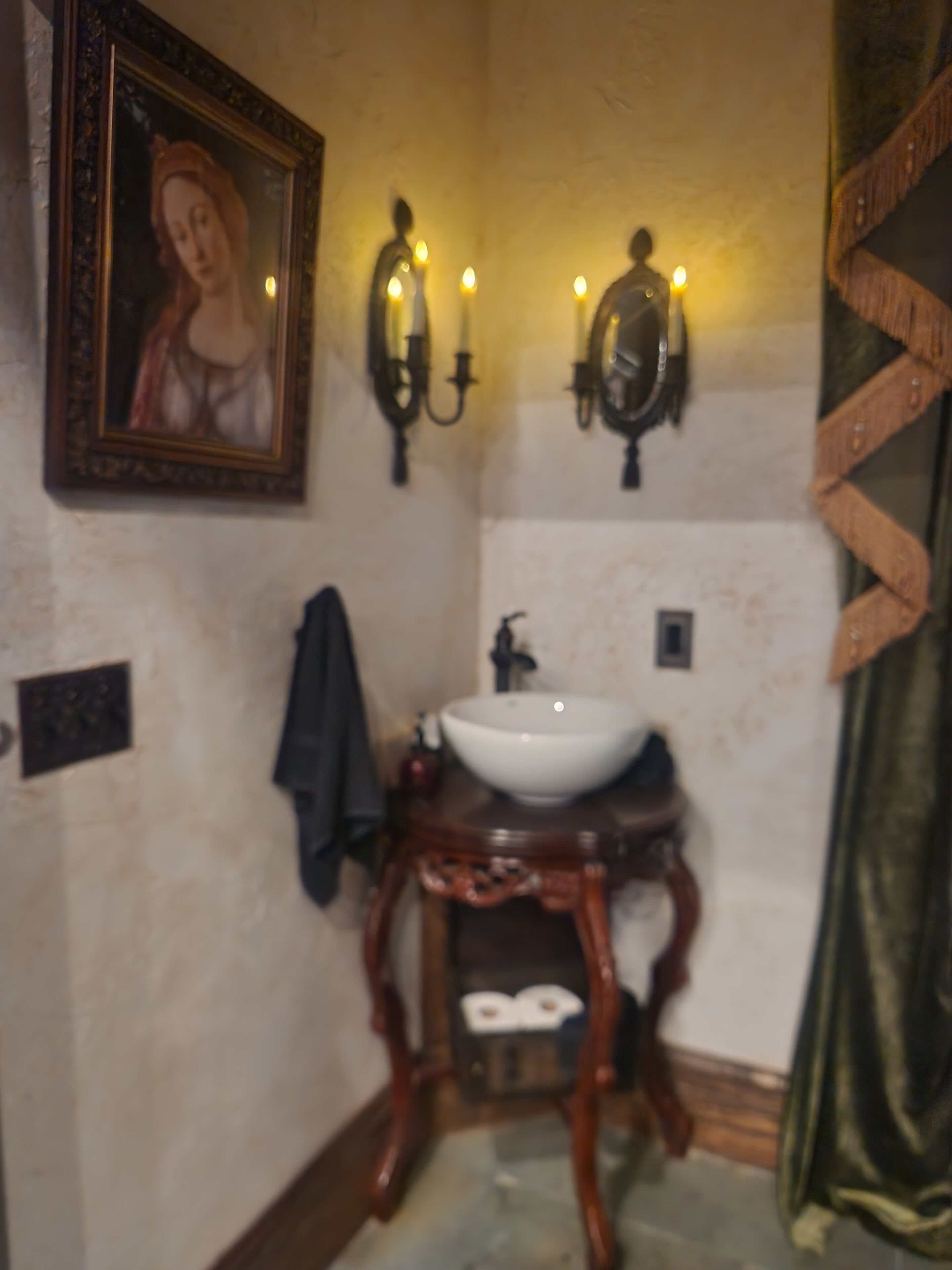 Un baño con un cuadro enmarcado, apliques de pared, un lavabo sobre un pedestal de madera y una cortina verde.