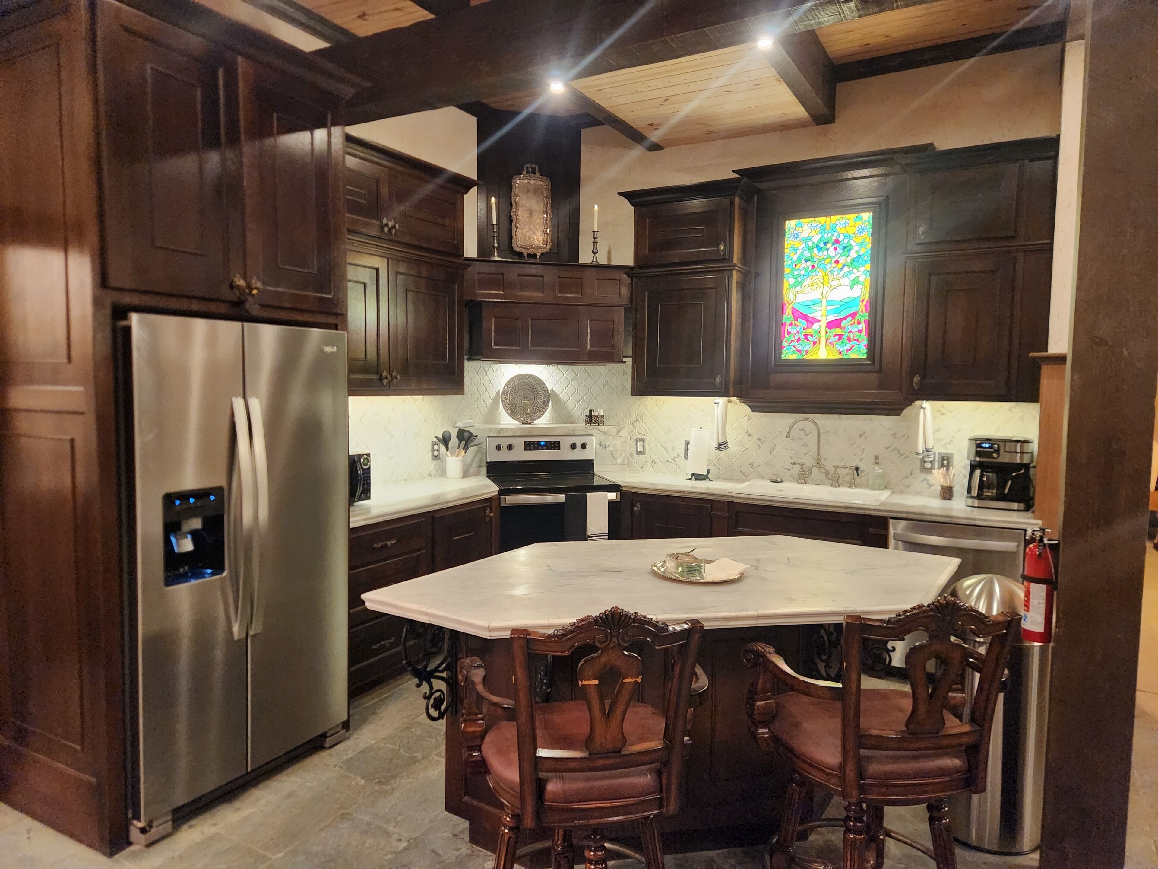 A kitchen with dark wooden cabinets, a stainless steel refrigerator, a large island with chairs, and a stained glass window.