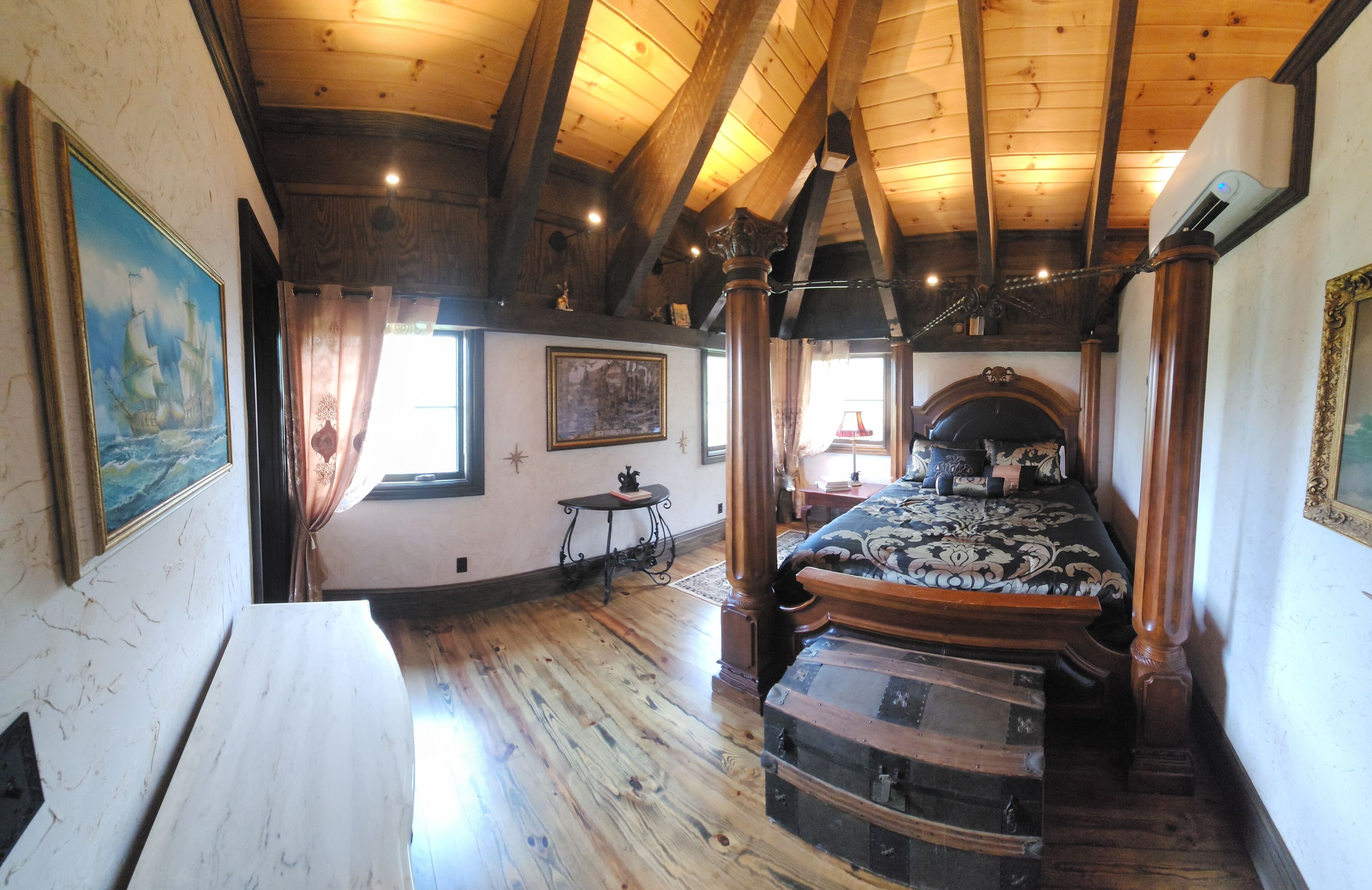 A room with a wooden ceiling, a bed with a patterned bedspread, a small table, and a painting on the wall.