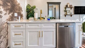 Cocina con gabinetes blancos, lavavajillas de acero inoxidable, un lavamanos con detalles dorados y un espejo grande sobre la encimera.
