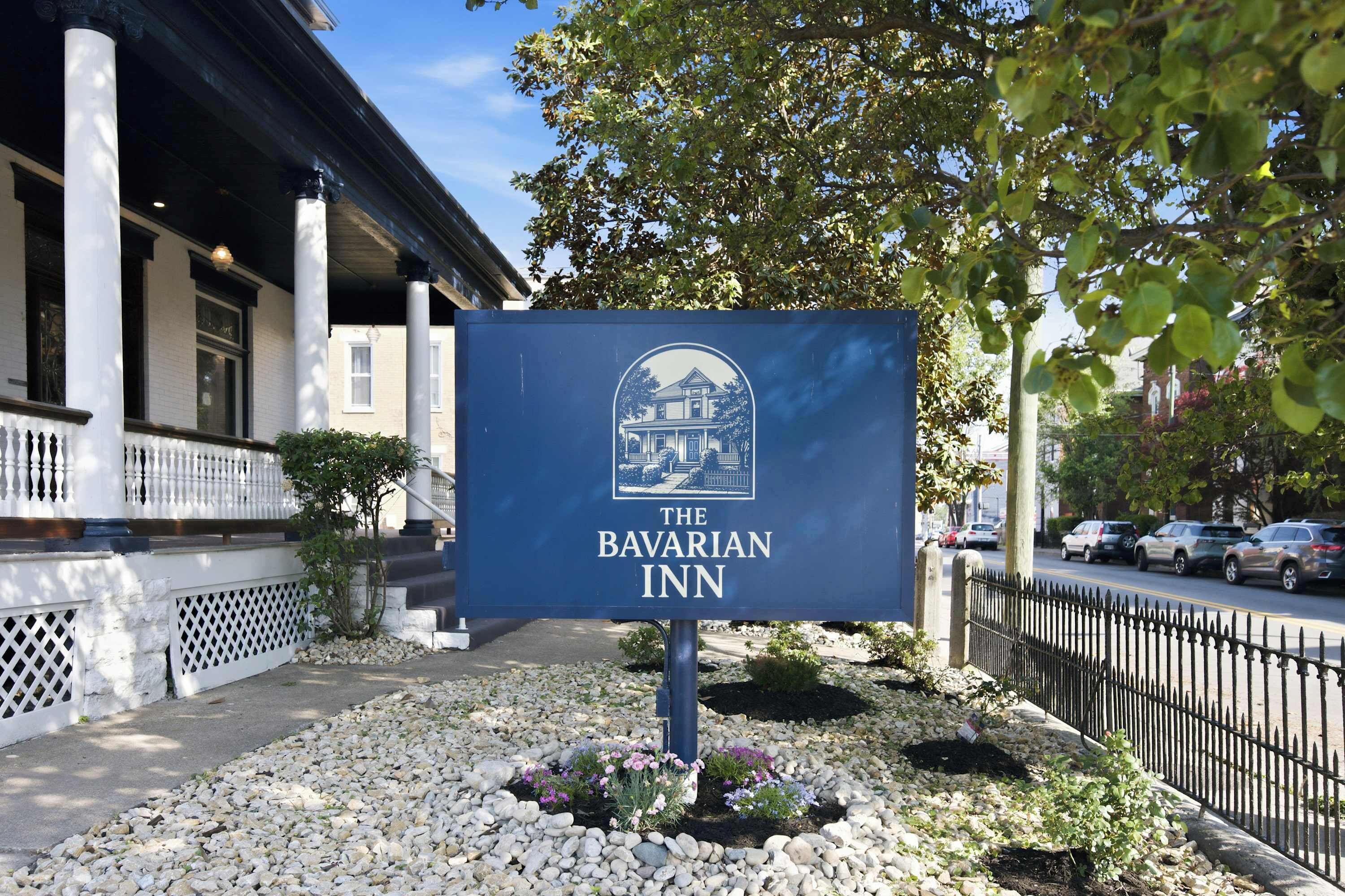 A sign for The Bavarian Inn with a building illustration, located in front of a building with columns and a fence.