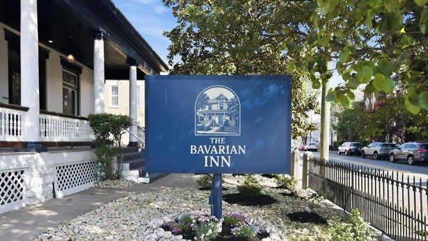 Un letrero para The Bavarian Inn con una ilustración del edificio, ubicado frente a un edificio con columnas y una cerca.