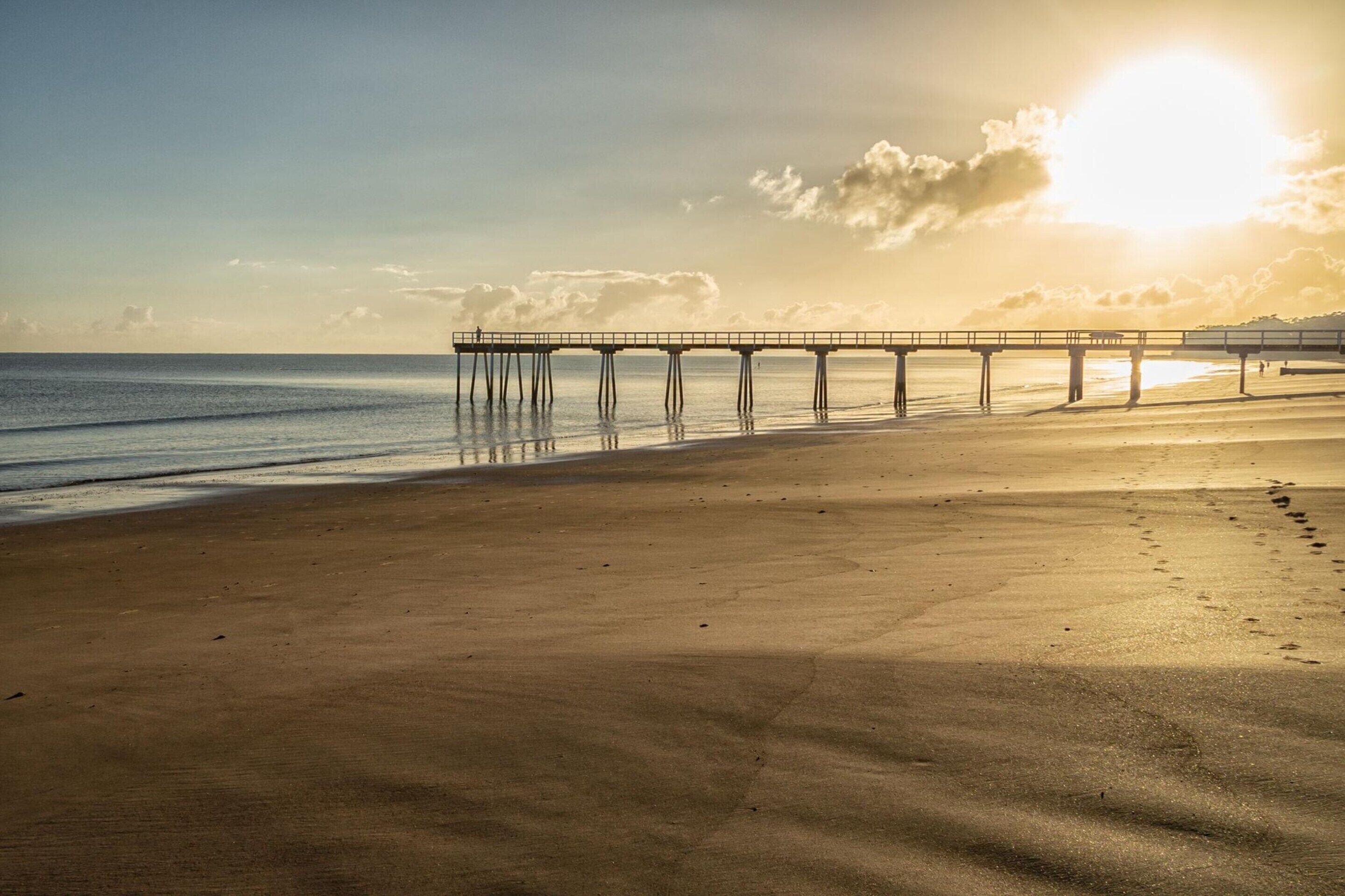 A beach with a pier extending into the sea, under a clear sky.
