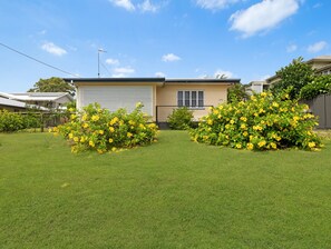 A single-story house with a garage, surrounded by well-maintained grass and yellow flowering bushes. - Two Bed Terrace Near Hervey Bay Beaches (Hervey Bay)