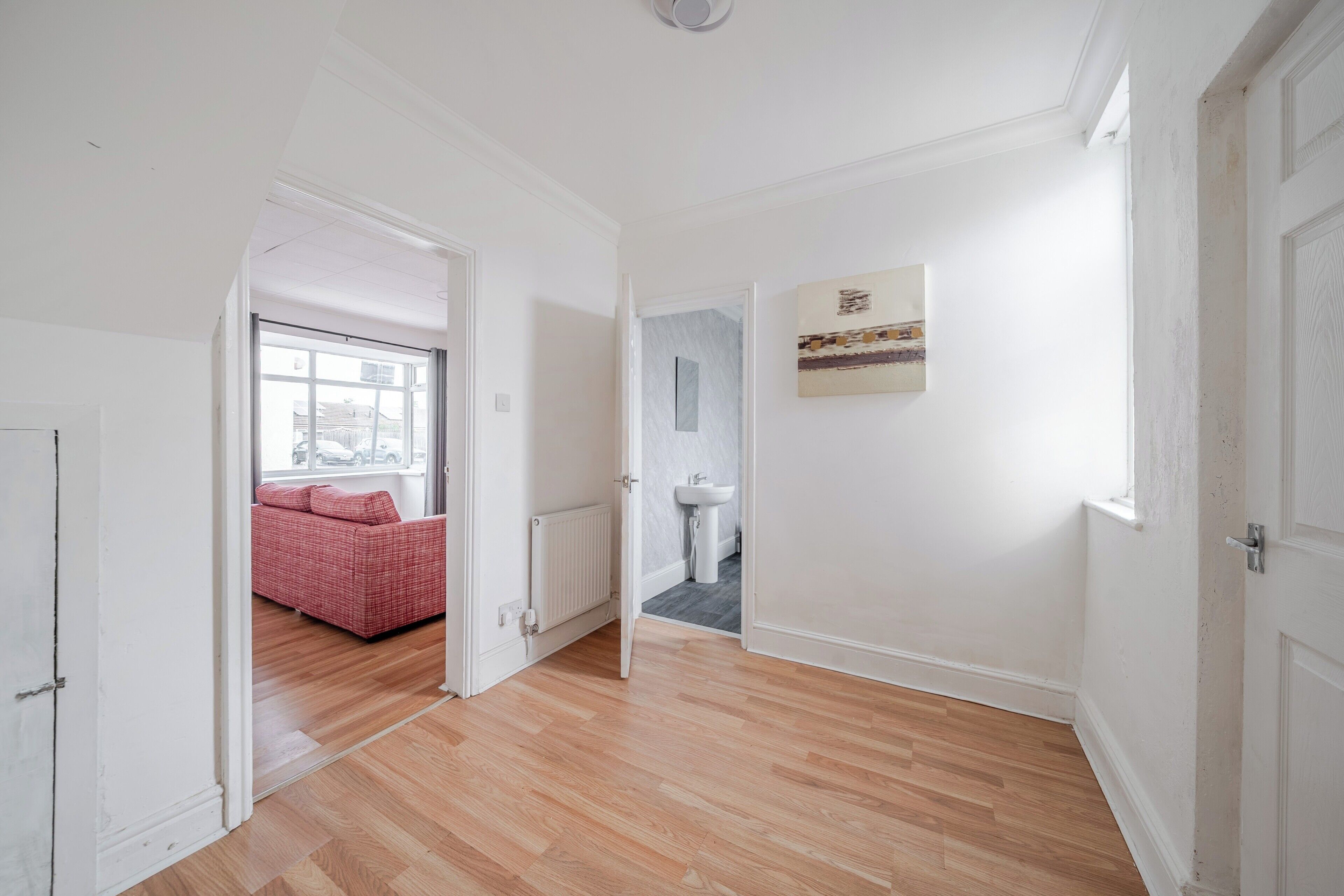 A hallway with a wooden floor, a red sofa in the background, a radiator, and a wall-mounted artwork.