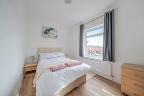 A bedroom with a bed, a nightstand, a dresser, a window with curtains, and a radiator.