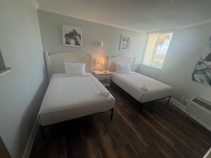 A room with two single beds, a nightstand with a lamp, a window with blinds, and a radiator. - Pelican Pointe 425:Walk to Beach~Pool~Free Parking (Clearwater Beach)