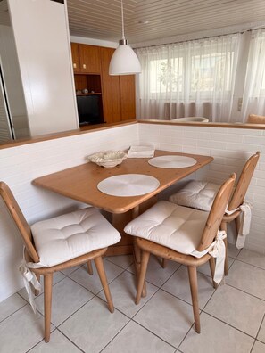 A dining area with a wooden table, four cushioned chairs, a white pendant light, and a kitchen in the background. - Rosmarie Vacation Rental—in the Heart of Rheinhessen (Alzey)