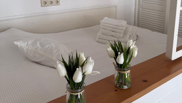 A bedroom with a bed, white pillows, a wooden bedside table, and two glass vases with white flowers. - Rosmarie Vacation Rental—in the Heart of Rheinhessen (Alzey)
