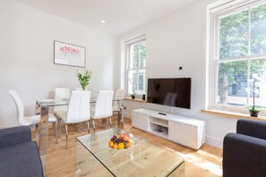 A modern living room with a glass coffee table, white furniture, a flat-screen TV, and large windows. - Modern Fitzrovia 3BR by Soho - London Centre (London)