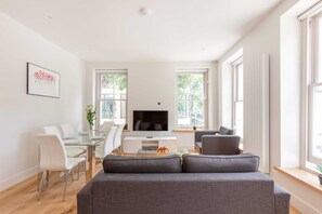Ein modernes Wohnzimmer mit einem Glastisch, weißen Stühlen, einem grauen Sofa und einem Flachbildfernseher.
