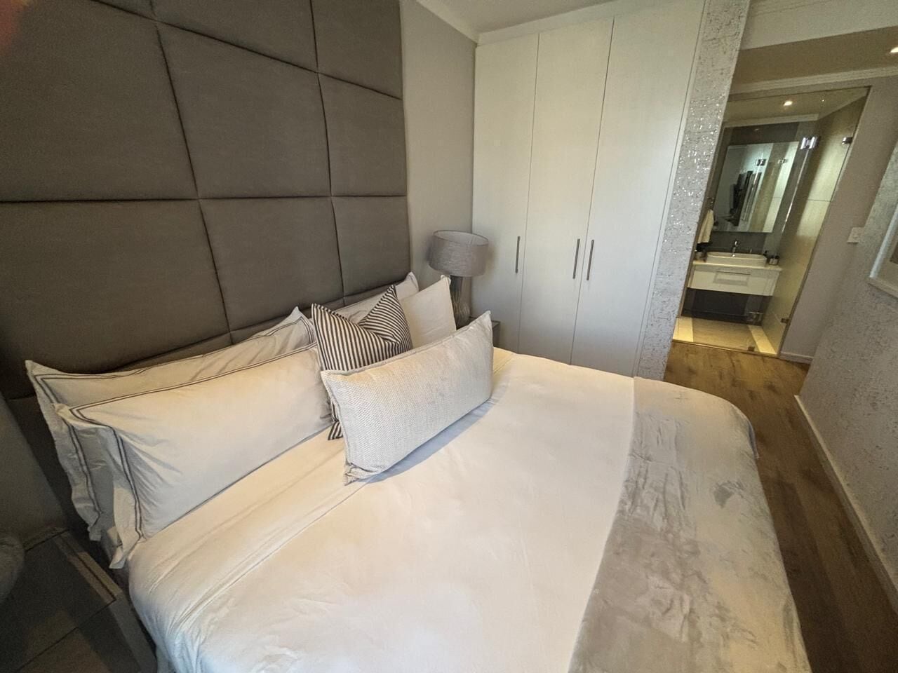 Un dormitorio con una cama grande, cabecera, mesita de noche con lámpara y armario.