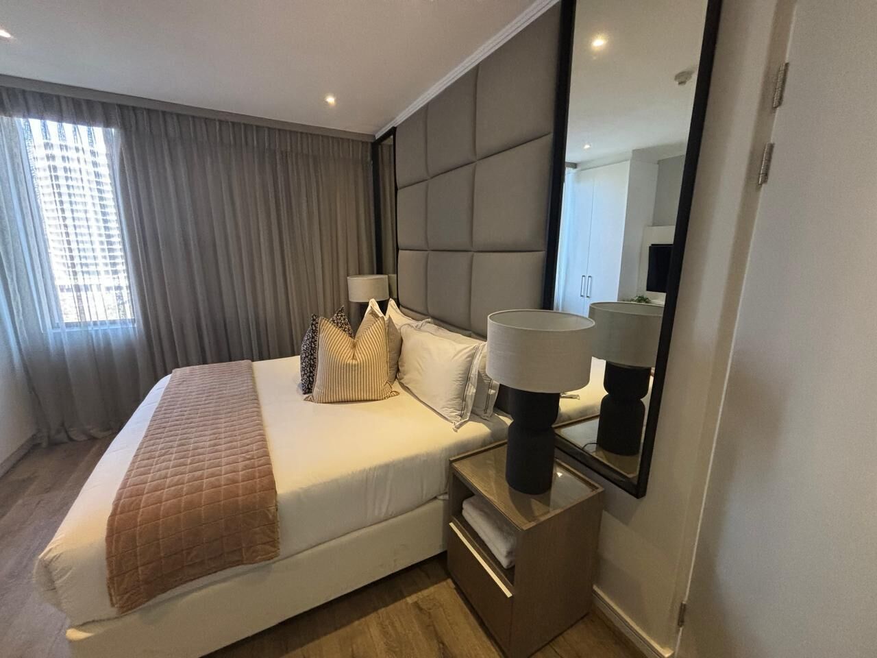 A hotel room with a bed, bedside lamps, a mirror, and a window with curtains.