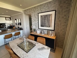 A modern kitchen with a marble countertop, a dining area with a glass-top table, and a framed artwork on the wall. -  Stunning 3-bedroom apartment in the heart of Sandton (Sandton)