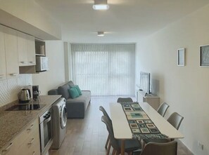 Una cocina moderna con lavadora integrada, una zona de comedor con una mesa larga y un espacio de sala de estar con sofá y televisor.