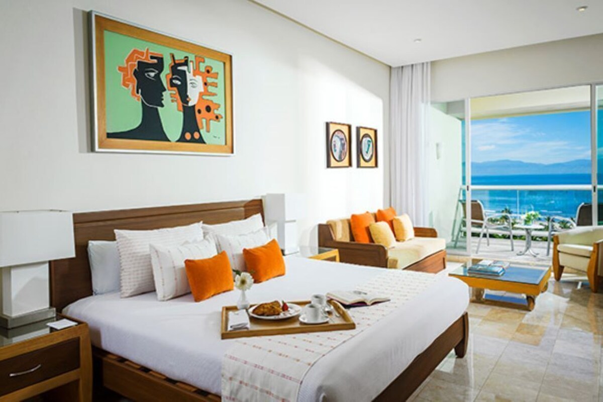A hotel room with a large bed, a desk, a chair, and a balcony with a view of the sea.