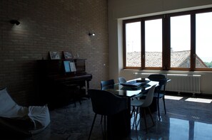 Une pièce avec un piano, une table à manger entourée de chaises, un radiateur et une grande fenêtre donnant sur un toit recouvert de tuiles.