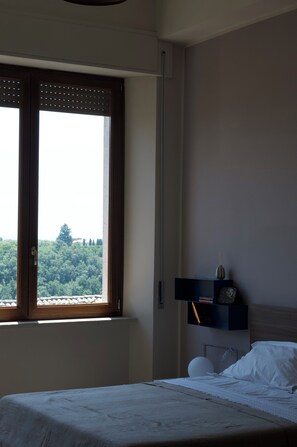 A bedroom with a bed, a nightstand, a window with blinds, and a view of trees and buildings.