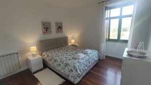 Ein Schlafzimmer mit einem Bett, zwei Nachttischlampen, einem Heizkörper, einem Fenster mit Vorhängen und einem kleinen Tisch mit Flasche und Gläsern.