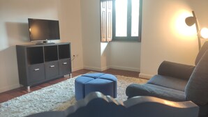 A modern living room with a flat-screen TV on a cabinet, a grey sofa, a blue ottoman, and a white shag rug. - Room in Ponte da Barca (Vila Nova de Muía)