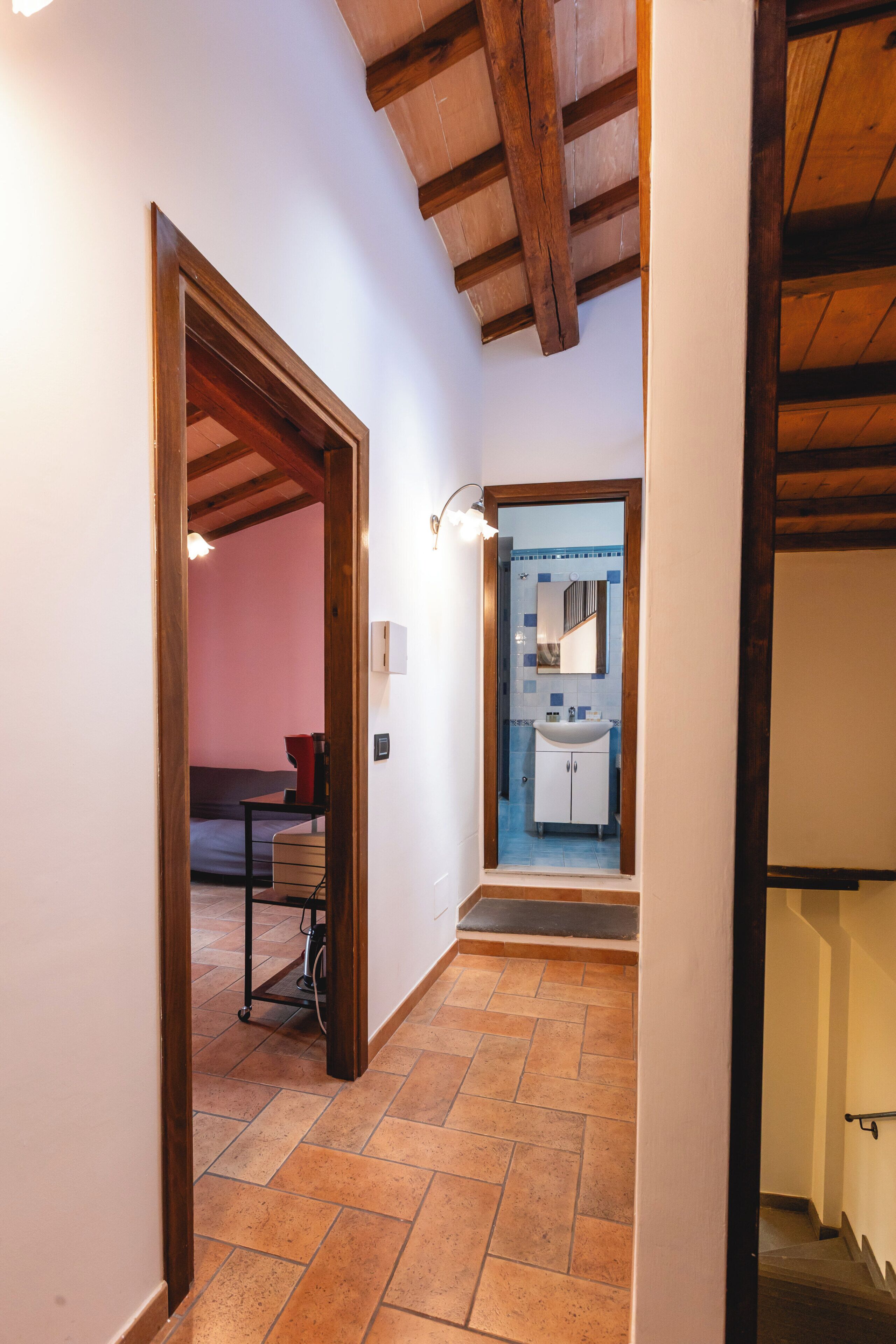 Un corridoio con pavimento piastrellato, travi in legno al soffitto, un lampadario e un bagno con lavandino e specchio.