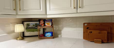 Eine Küche mit weißen Schränken, einer Marmorarbeitsplatte, einem Korbschrank mit Snacks, einem gerahmten Bild und einem Holzschneidebrett.