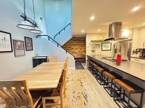 Eine moderne Küche mit einer Holztreppe, einem Essbereich mit einem langen Tisch und Stühlen sowie einer Bar mit Hockern.