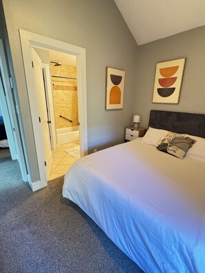 Ein Schlafzimmer mit einem Bett, einem Nachttisch, einem Wandbild und einer offenen Tür zu einem Badezimmer mit Dusche und Badewanne.