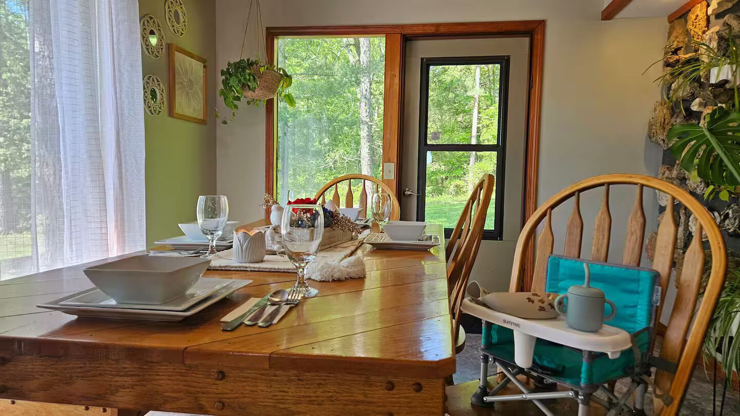Een eetruimte met een houten tafel voor twee personen, stoelen en een uitzicht op de omgeving door grote ramen.