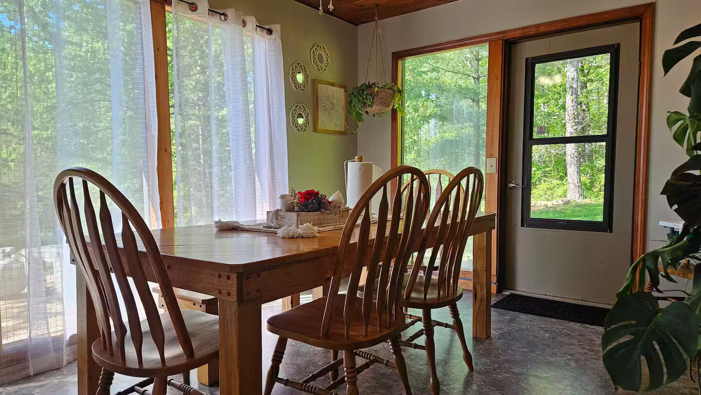 A wooden dining table with four chairs, a window with sheer curtains, and a door with a view of greenery.