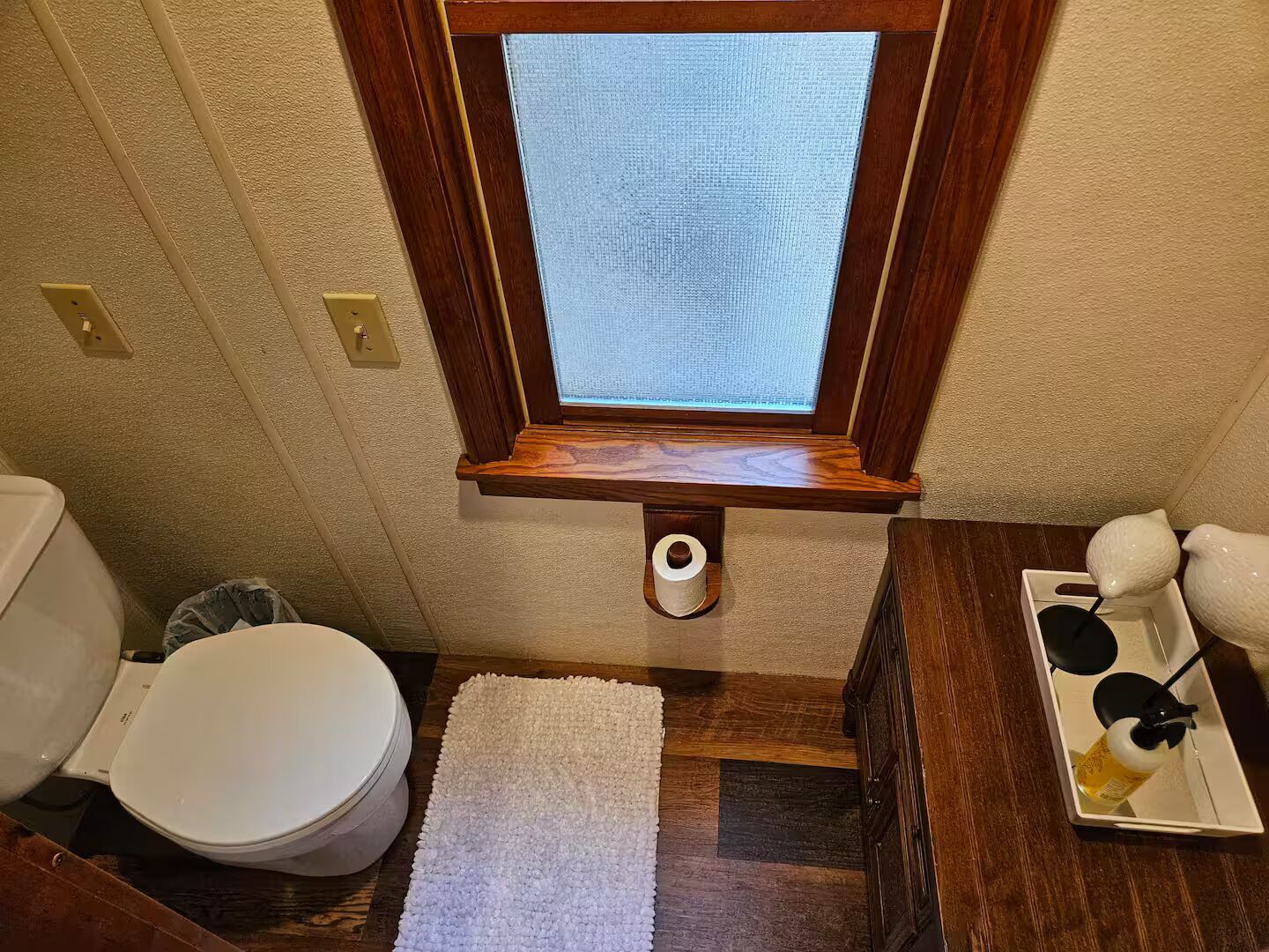 Un baño pequeño con inodoro, ventana, estante y alfombra blanca.