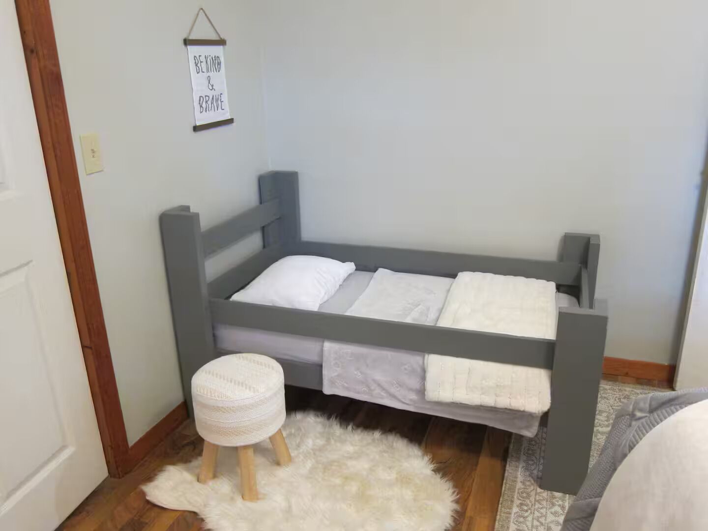 A single bed with a grey frame, a white pillow, and a white blanket. There is a small stool with a white cushion and a fluffy rug on the floor. A sign with the words 'BE KIND & BRAVE' hangs on the wall.
