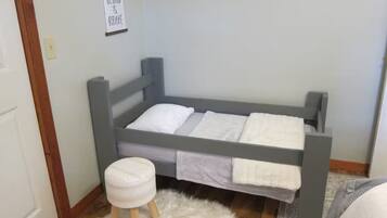 A single bed with a grey frame, a white pillow, and a white blanket. There is a small stool with a white cushion and a fluffy rug on the floor. A sign with the words 'BE KIND & BRAVE' hangs on the wall.