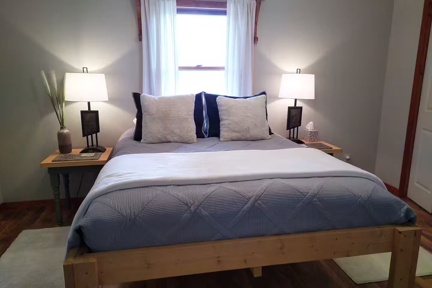 Un dormitorio con una cama de madera, dos mesitas de noche con lámparas, una ventana con cortinas y un mueble auxiliar con una caja de pañuelos.