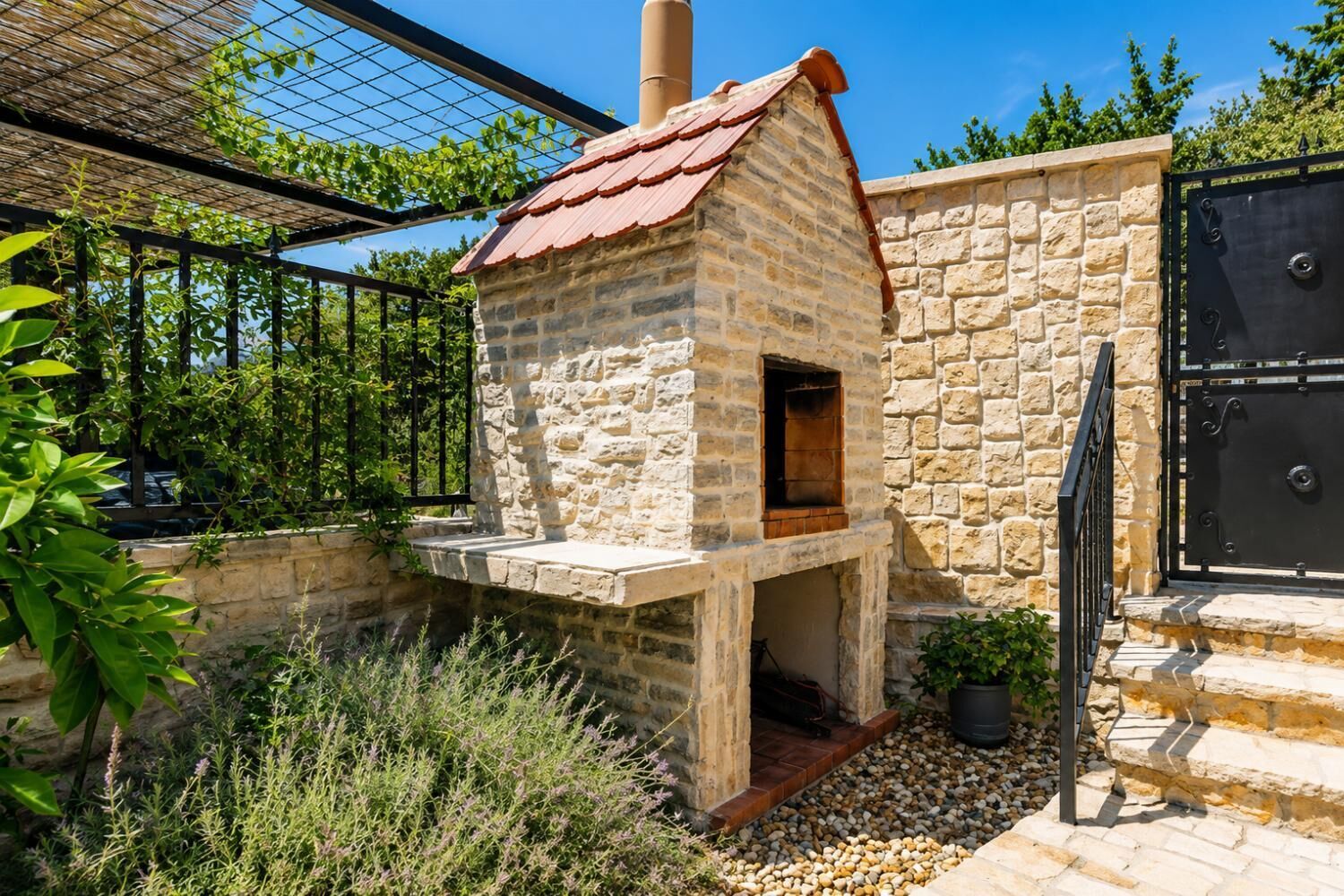 A stone-built outdoor oven with a red roof, surrounded by greenery and a stone wall.