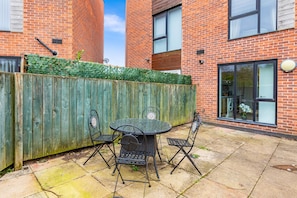 A patio with a round table and four chairs, a wooden fence, and a brick building with windows. - Cozy, modern home in the heart of Manchester, close to the city center. (Salford)
