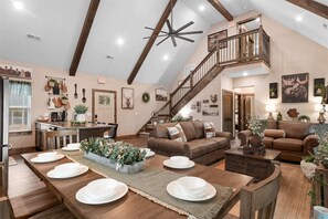 A spacious living area with a high ceiling, a staircase, and a dining setup with white plates. - The Lazy Beaver Lodge Hot Tub (Hollister)