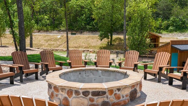 A circular stone fire pit surrounded by wooden Adirondack chairs, set on a gravel area with trees in the background. - The Lazy Beaver Lodge Hot Tub (Hollister)