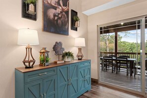 A room with a teal cabinet, two wall-mounted lamps, a moose painting, and a view of an outdoor deck with tables and chairs. - The Lazy Beaver Lodge Hot Tub (Hollister)