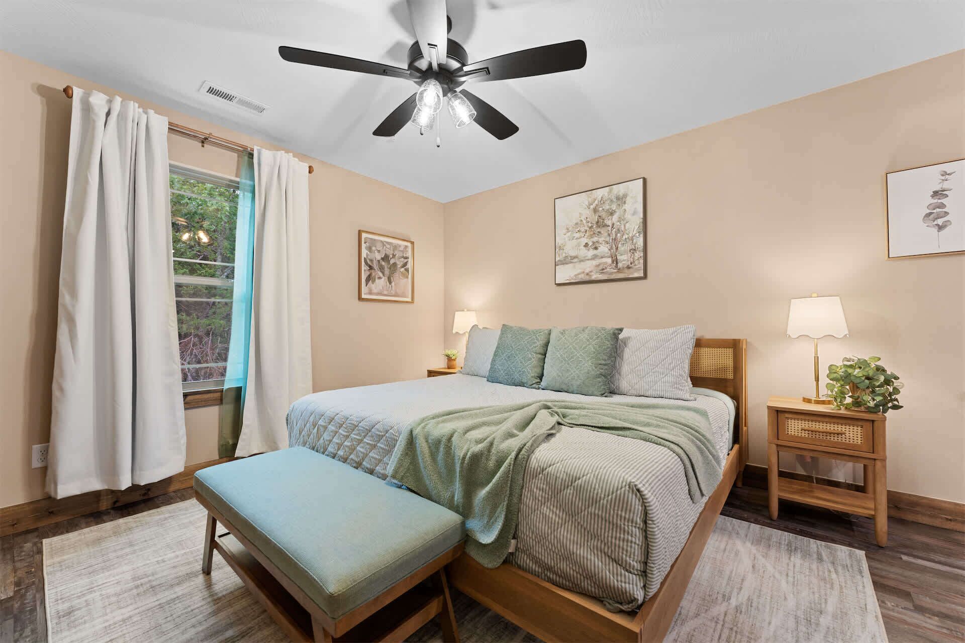 A bedroom with a bed, a bench, a ceiling fan, a window with curtains, and a nightstand with a lamp and a plant.