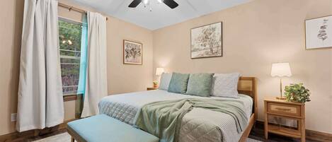 A bedroom with a bed, a bench, a ceiling fan, a window with curtains, and a nightstand with a lamp and a plant.