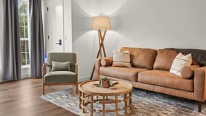 Ein Wohnzimmer mit einem Ledersofa, einem Holztisch, einem grünen Sessel, einer Stehlampe und einem gemusterten Teppich.