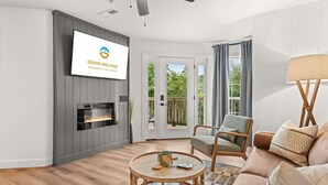 Ein modernes Wohnzimmer mit Kamin, Wand-Fernseher, Deckenventilator und Blick ins Freie durch eine Glastür.