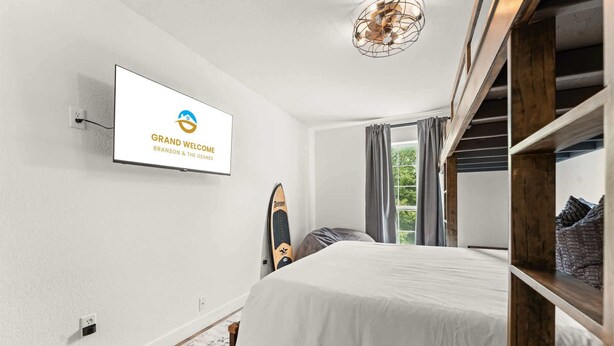 Ein Hotelzimmer mit Bett, einem an der Wand befestigten Fernseher, der das Logo des Hotels zeigt, einem Surfbrett und einem Fenster mit Vorhängen.